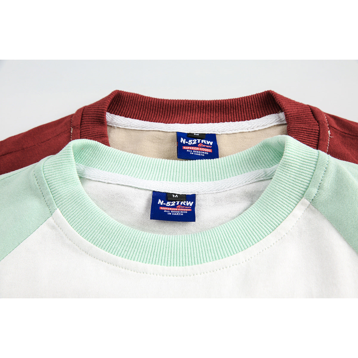 Close-up of overlapping white and beige raglan shirts with mint and burgundy ribbed collars and sewn neck labels.