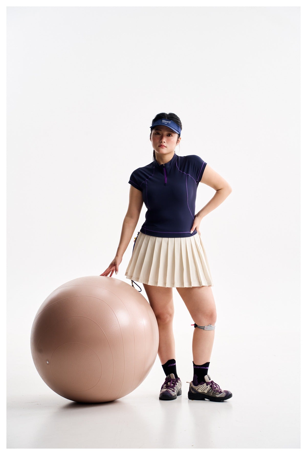 Model wearing a navy zip-neck top with purple piping and a cream pleated skirt, beside a large exercise ball.
