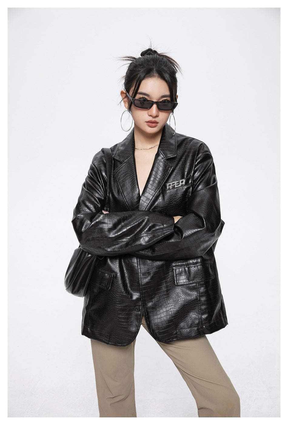 Model with crossed arms wearing the black crocodile-embossed oversized jacket with khaki pants and a black shoulder bag