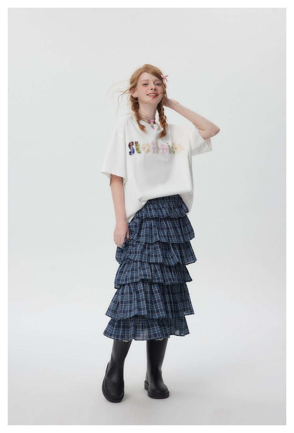 Model wearing a white appliqué-letter T-shirt with a blue plaid tiered skirt and black boots, full-length view