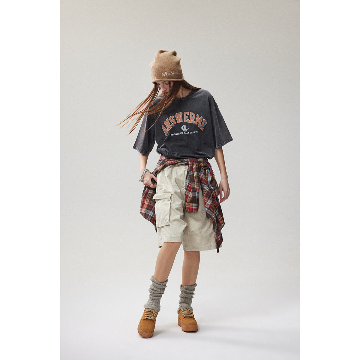 Full-length model wearing a charcoal graphic T-shirt, beige cargo shorts, and a red plaid shirt tied at the waist.