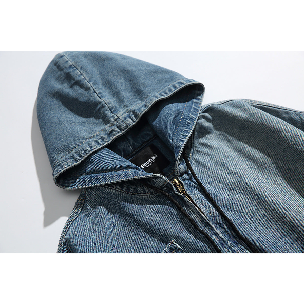 Close-up of a blue washed-denim jacket showing its hood, black drawstrings, brass zipper and sewn-in neck label.