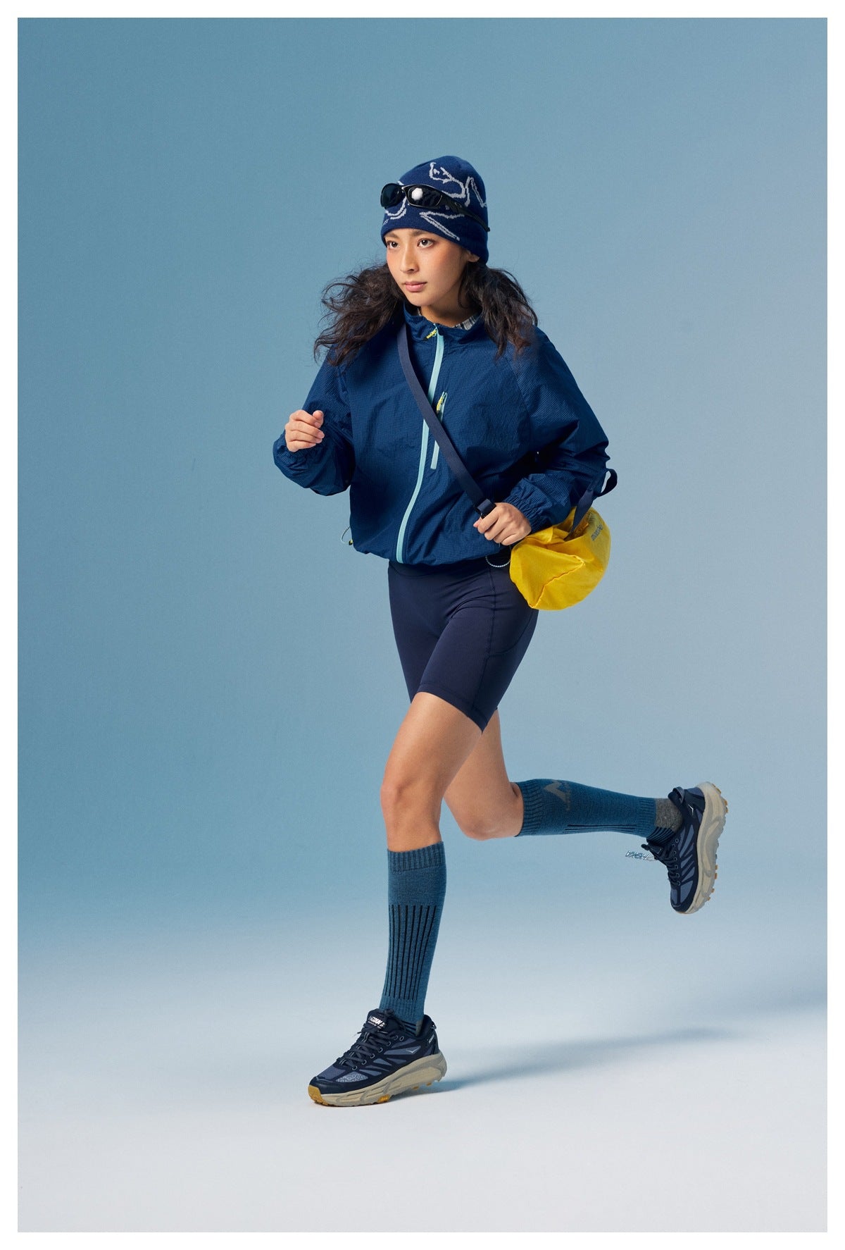 Model in a navy zip-front jacket and dark shorts, shown in motion with blue knee socks and a yellow shoulder bag.