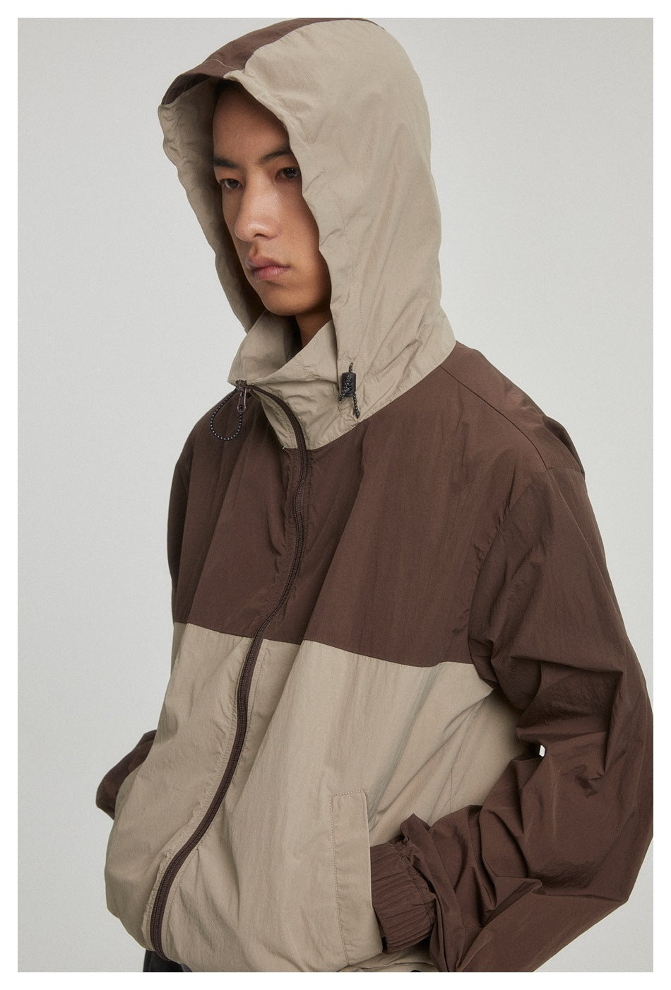 Close view of a model wearing a brown-and-beige color-block jacket with the beige hood raised and front zip visible.