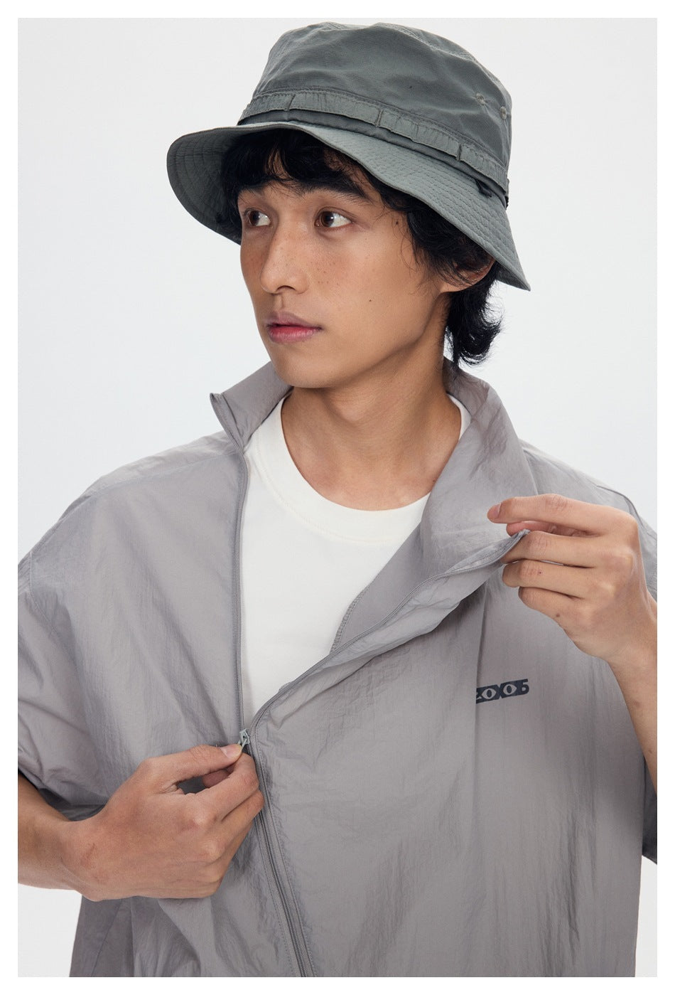 Close-up of a model wearing a light gray zip-front short-sleeve jacket and dark gray bucket hat.