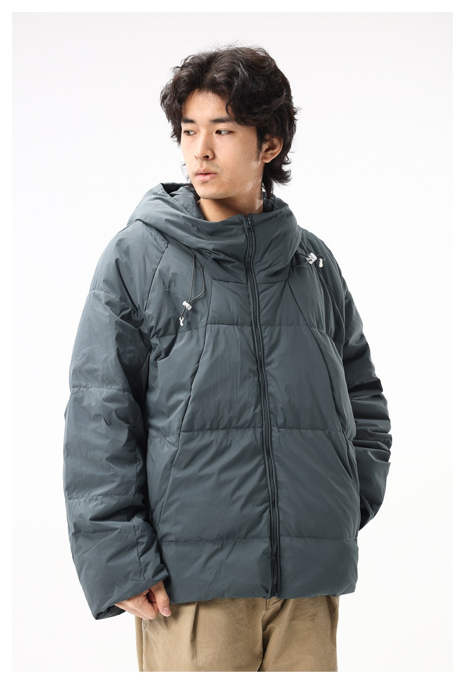Model wearing a slate gray hooded puffer jacket, shown from a slight angle with its front zipper and drawcords visible.
