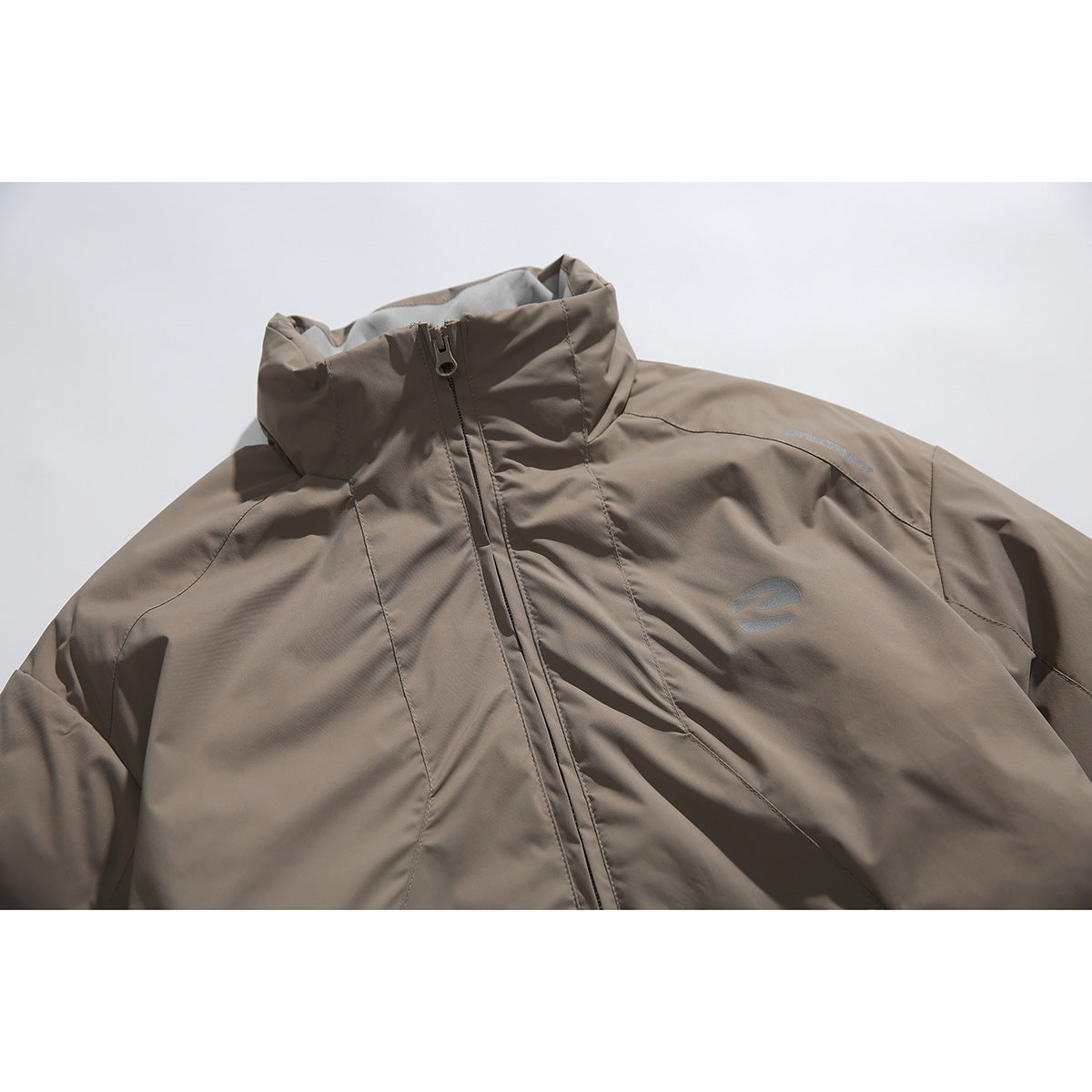 Close-up of a taupe jacket showing its raised collar, front zipper, chest emblem and small shoulder lettering.