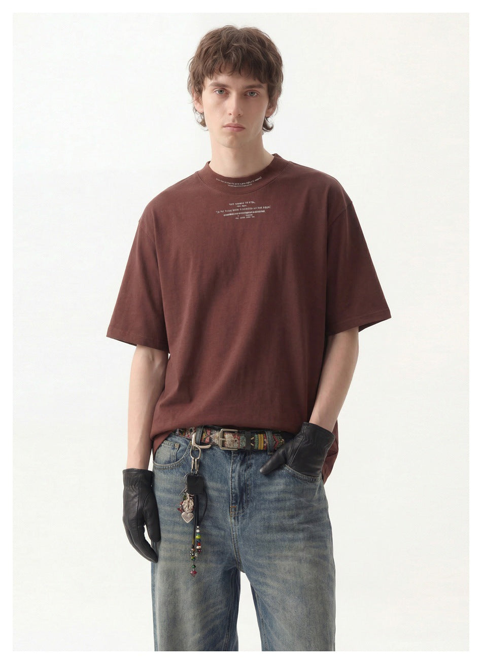 Man wearing a reddish-brown loose T-shirt with chest text print, faded jeans and black leather gloves, close-up view