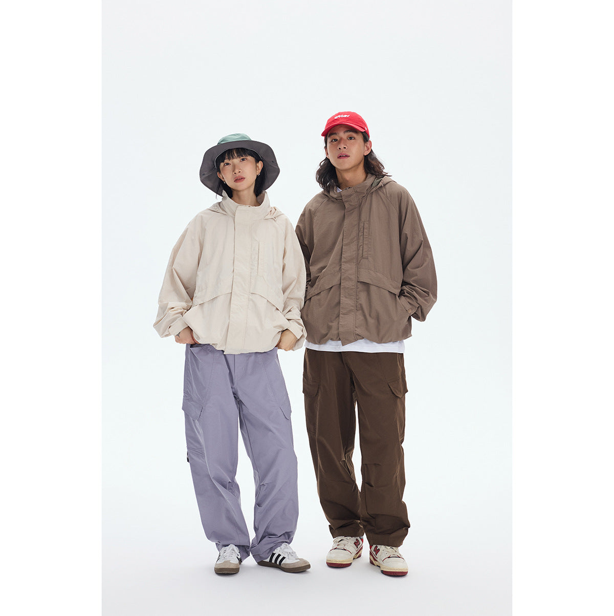 Two models wear hooded jackets in cream and taupe with loose cargo pants and brimmed hats.