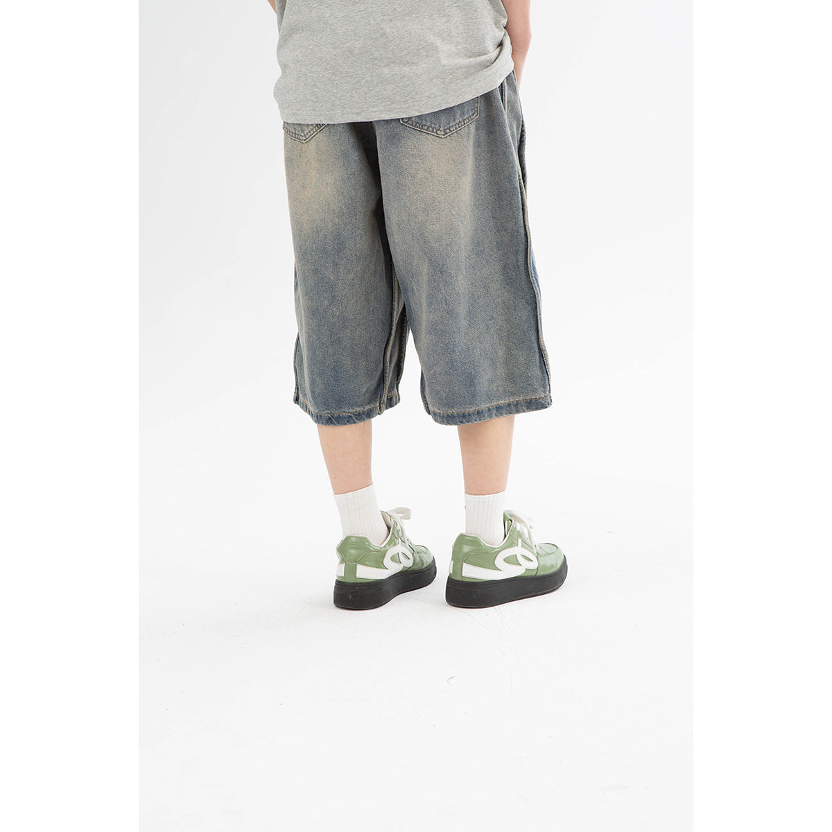 Rear view of loose, washed blue-gray denim shorts with two back pockets, worn with white socks and green sneakers.