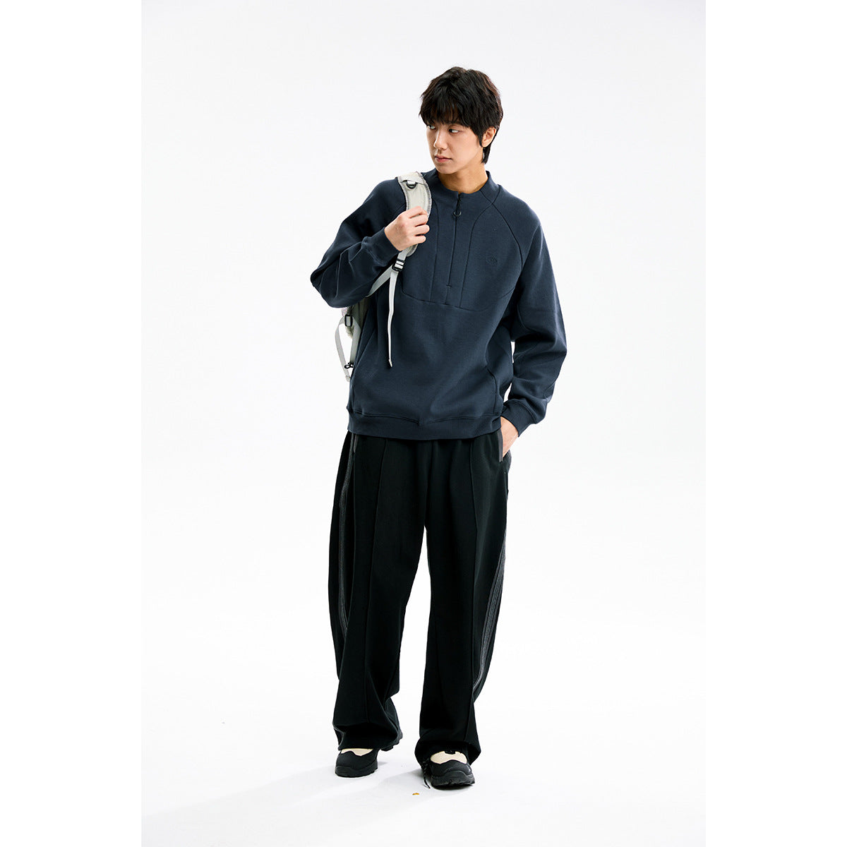Model wearing a dark navy zip-neck pullover and black wide-leg pants, with a light backpack over one shoulder.