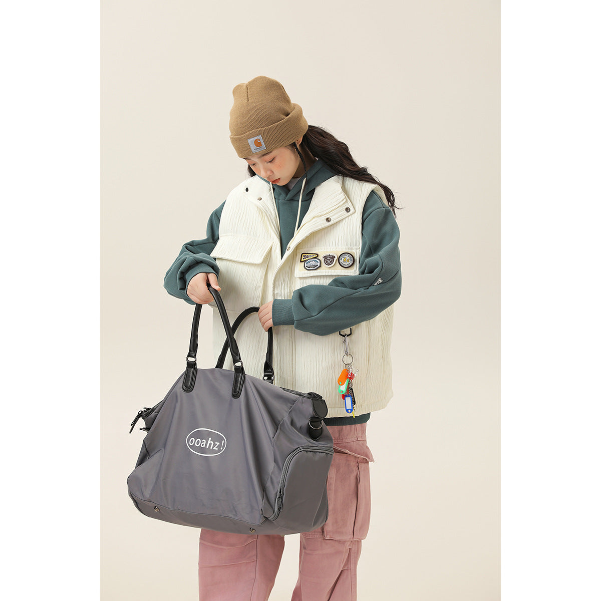 Model in an ivory utility vest over a teal hoodie and pink cargo pants, holding a large gray tote.