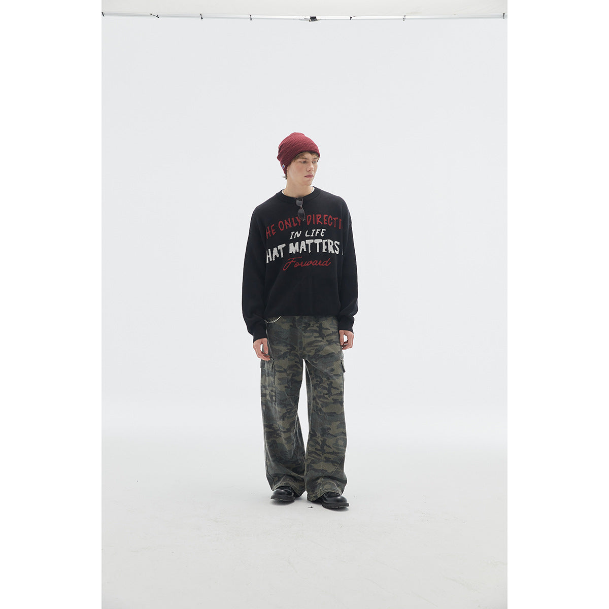 Model wearing a black slogan sweatshirt, red knit beanie, and loose green camouflage cargo pants.