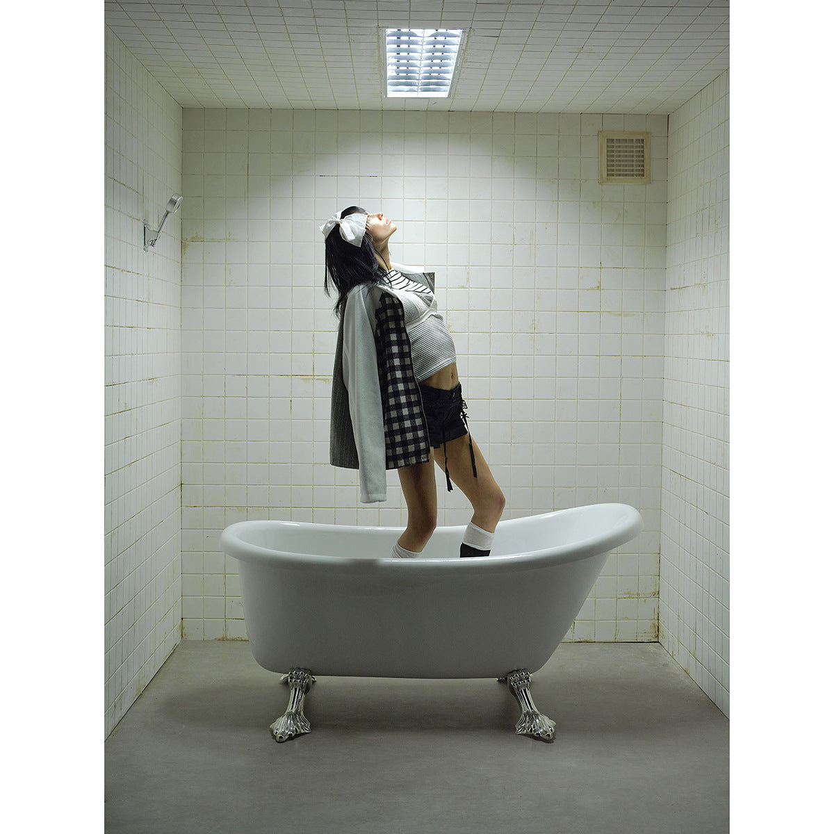 Model in a patchwork jacket leans back in a bathtub; the jacket has white sleeves and check, gray and dark panels.