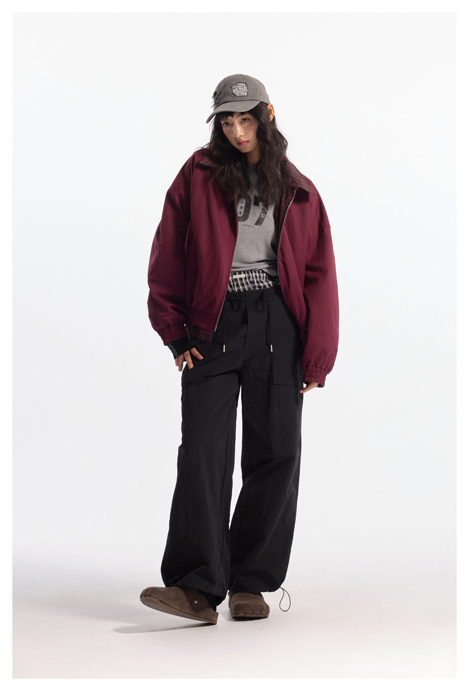 Model in burgundy zip jacket with plaid lining visible, gray tee, black wide-leg pants and cap