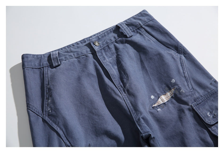 Close-up of blue-gray cargo pants showing the waistband, fly, pocket seams, and a distressed opening with plaid fabric