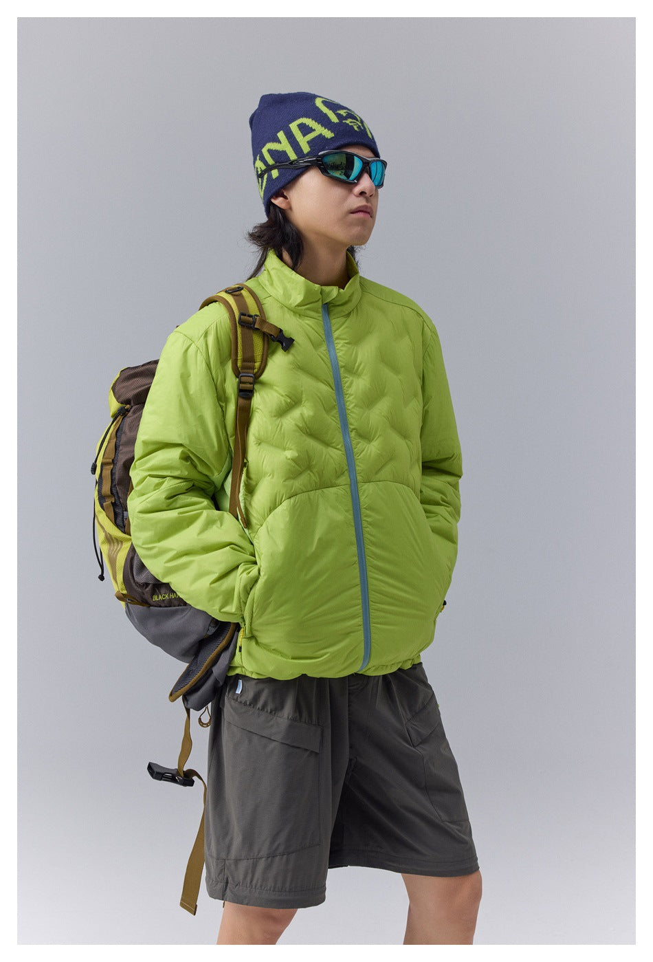 Model wears a lime green quilted jacket with blue zipper trim and charcoal shorts, carrying a hiking backpack.
