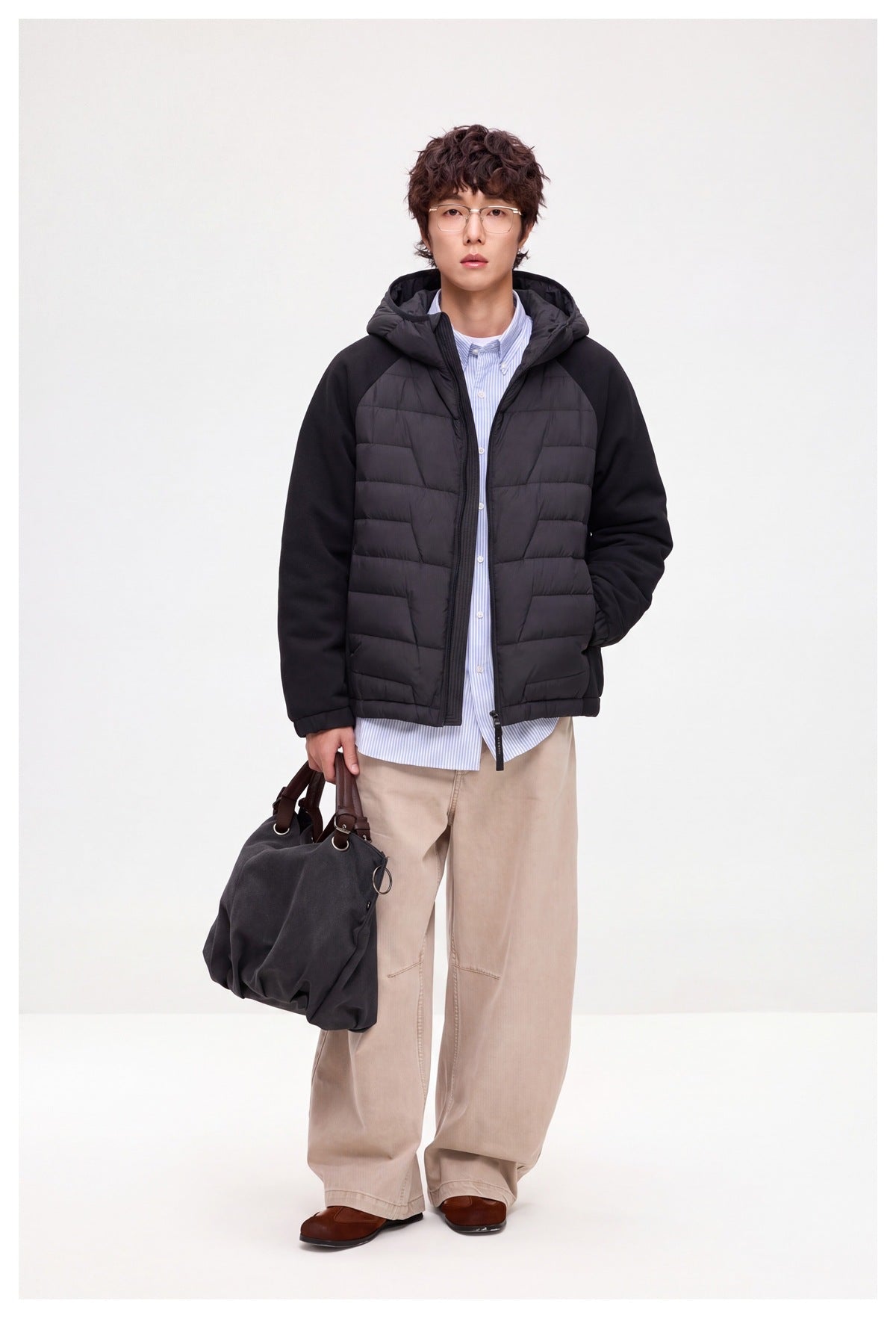 Man wearing black quilted hooded zip jacket over a striped shirt with beige wide-leg pants, holding a dark tote bag