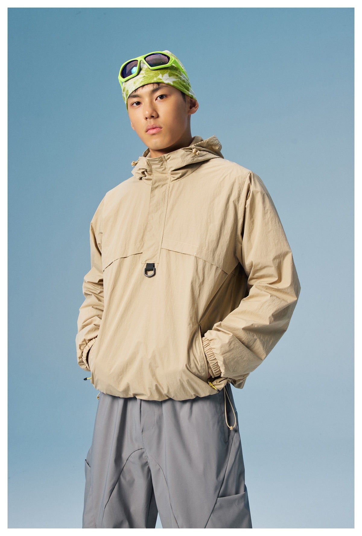 Model wearing a beige hooded pullover jacket with a front flap and gray trousers.