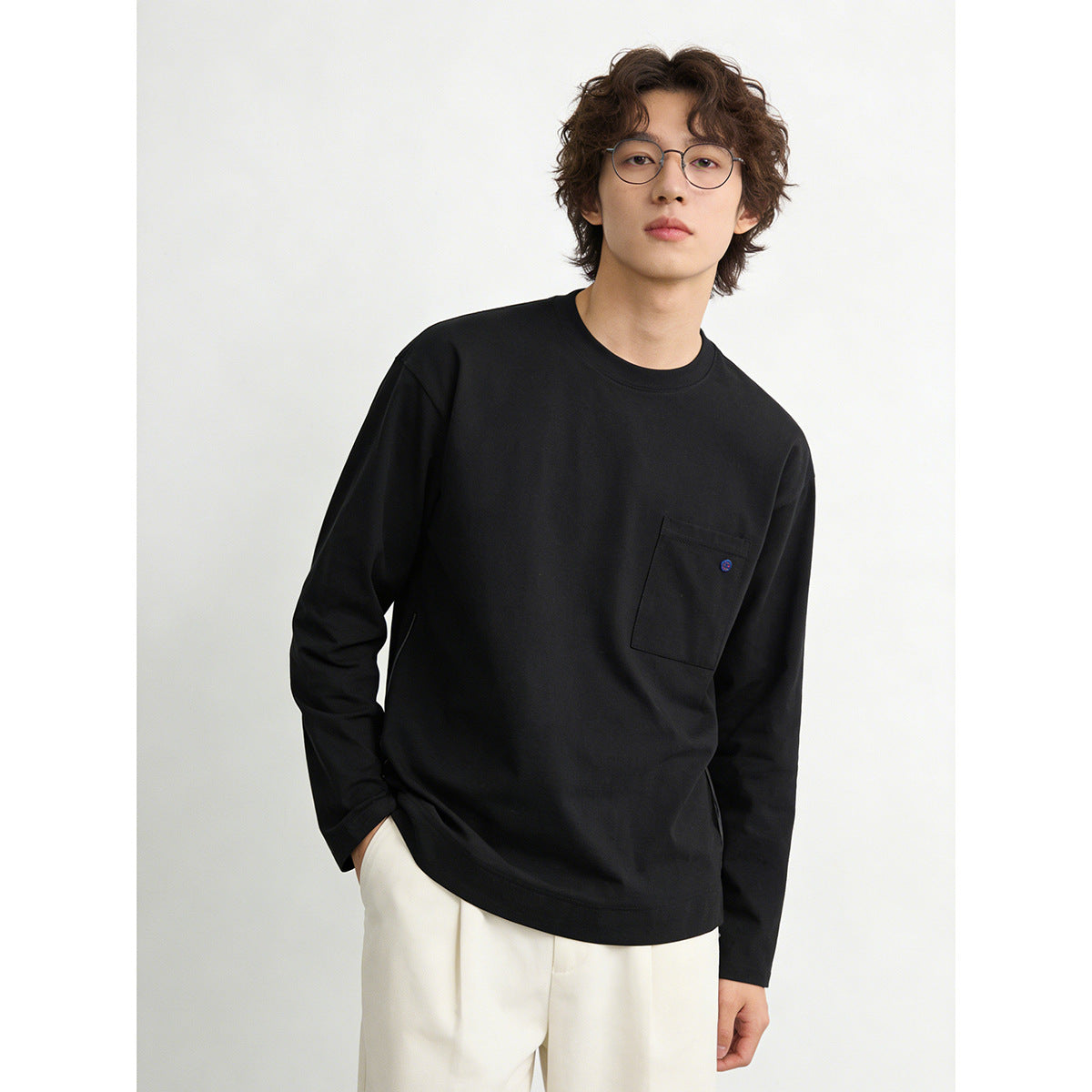Closer view of a model wearing a black long-sleeve crewneck shirt with a chest pocket and white trousers.