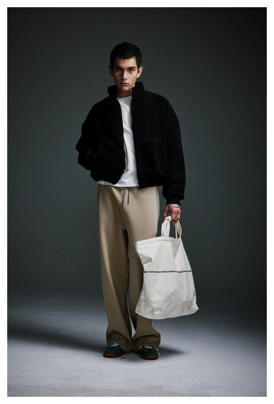 Model wearing the black fleece-like zip jacket with one hand in pocket, with white tee, beige pants and a canvas tote