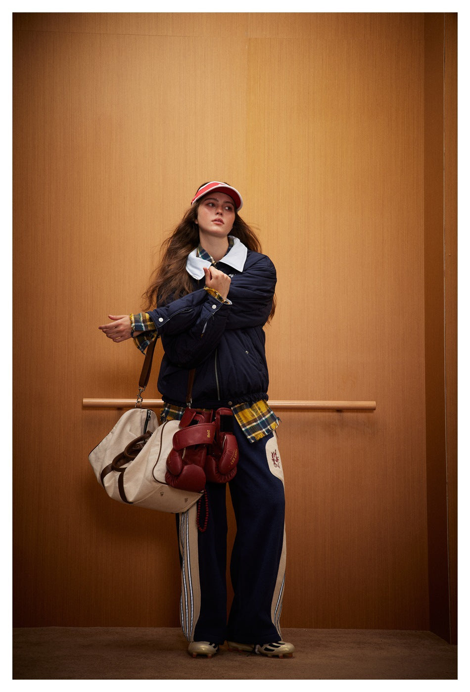Model wearing navy side of the zip jacket over a plaid shirt with wide-leg trousers, visor, duffel bag and boxing gloves