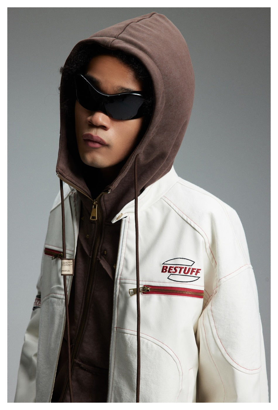 Close-up of a model wearing a white jacket with red trim over a zipped brown hood and black sunglasses.