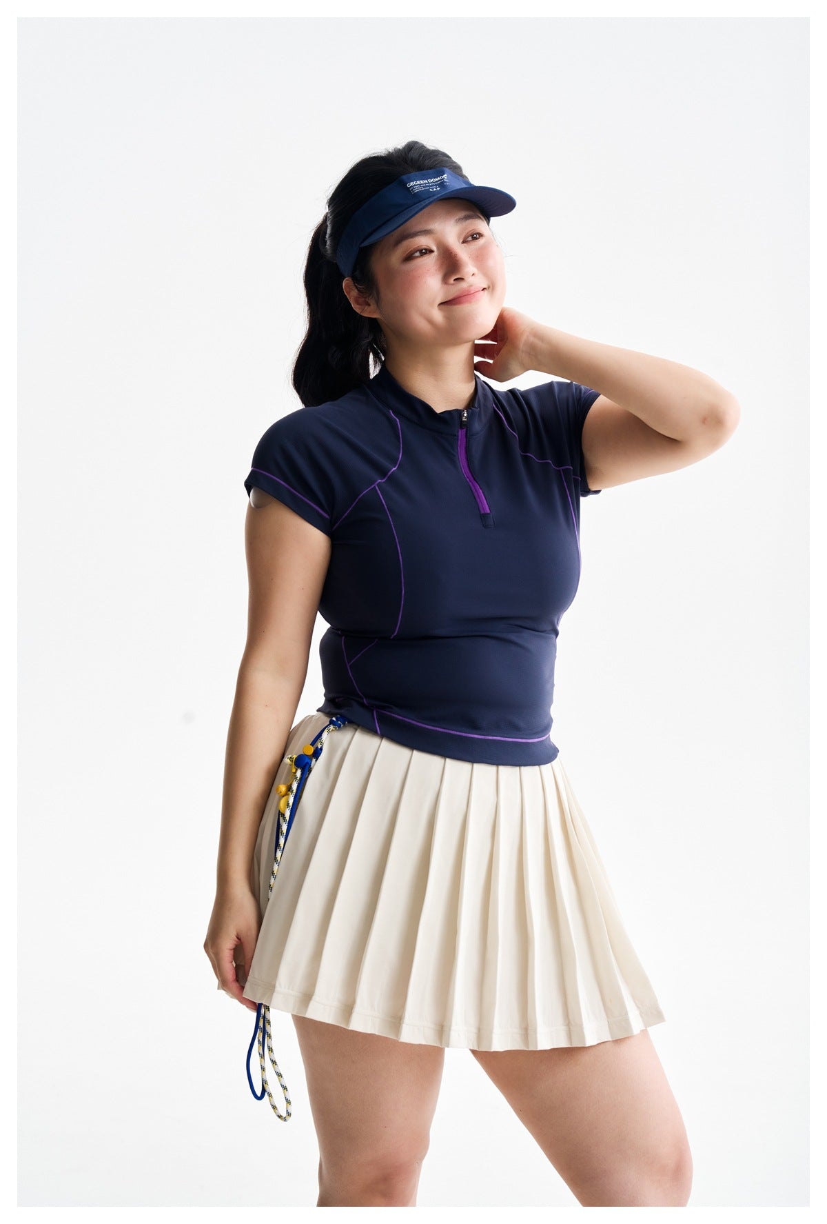 Model in a navy zip-neck sports top with purple seam accents and a cream pleated skirt, shown at an angle.