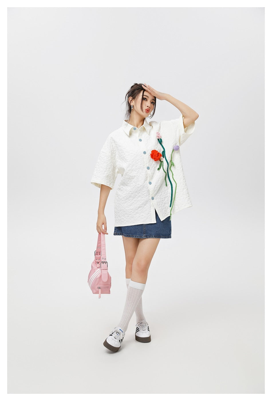 Model wearing a white textured short-sleeve shirt with colorful flower appliqués, paired with a denim skirt.