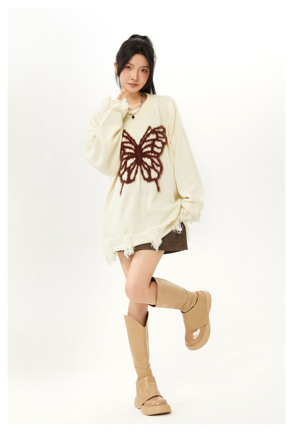Model posing in an oversized ivory butterfly sweater with frayed cuffs and hem, a brown skirt, and tan boots.