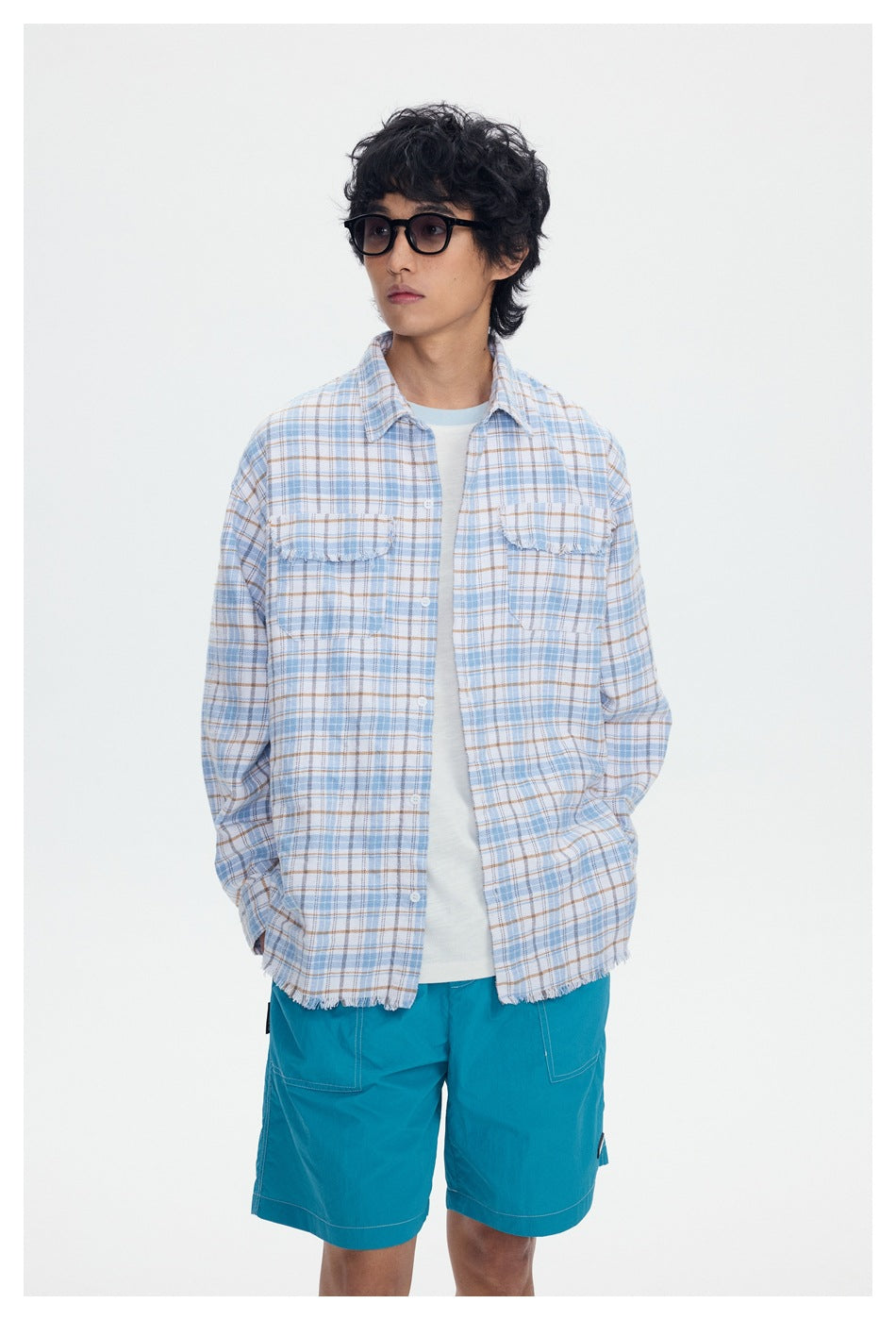 Model wearing a light blue and tan plaid shirt with frayed edges over a white tee and turquoise shorts, front view.