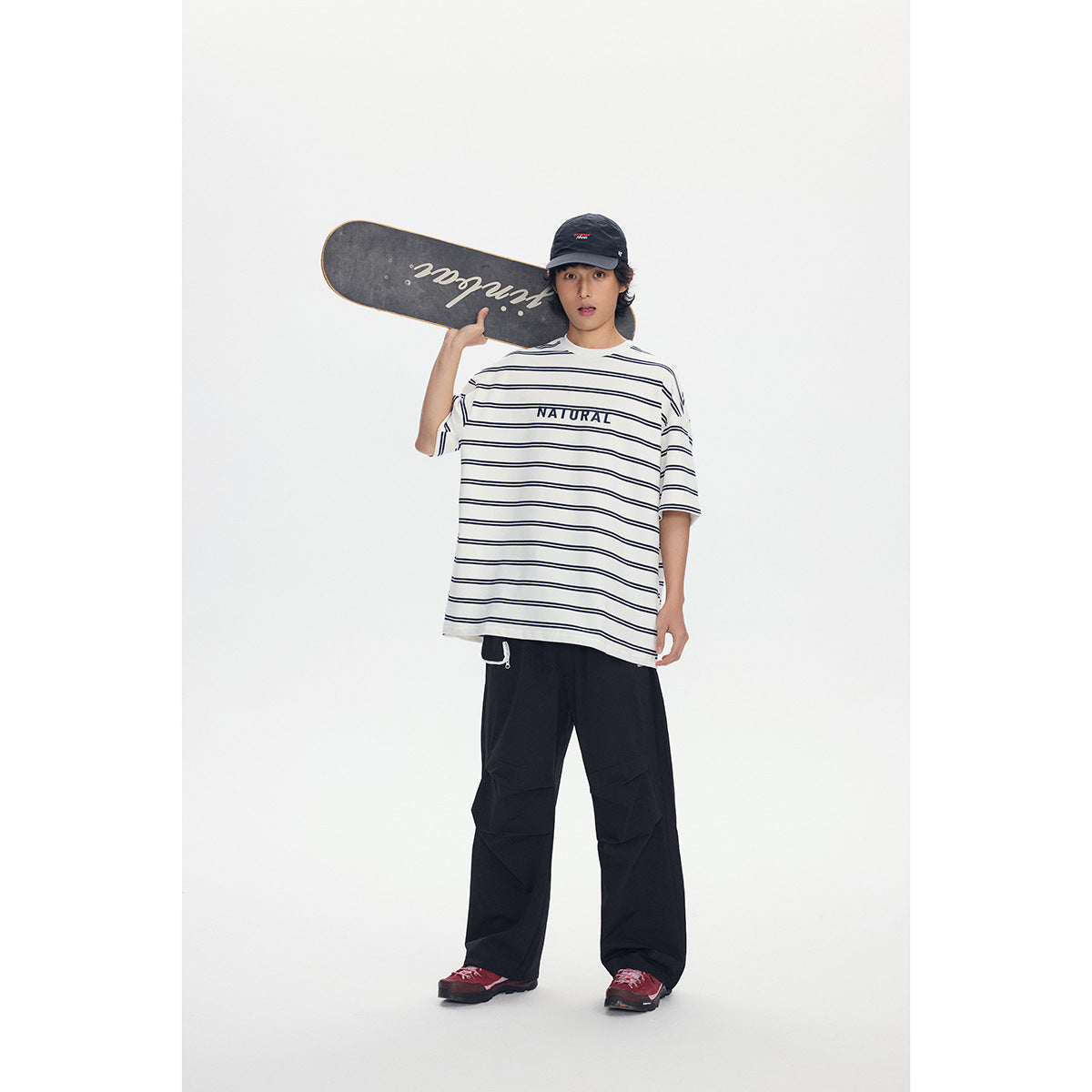 Full-length model wearing an ivory T-shirt with navy stripes and NATURAL lettering, black pants and a cap, holding a