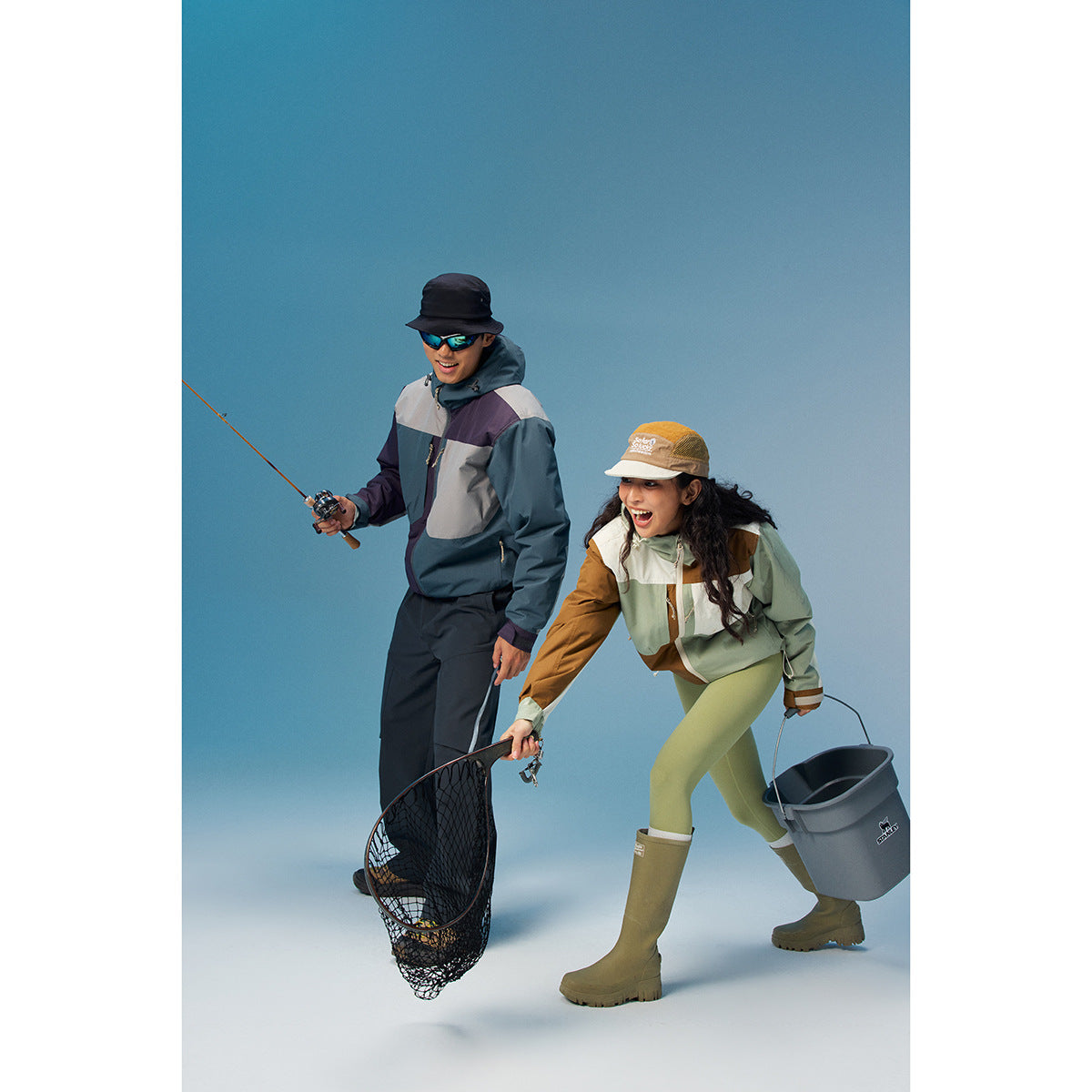 Two models fishing in color-block hooded jackets; one wears sage, cream, and brown, the other blue, gray, and purple.