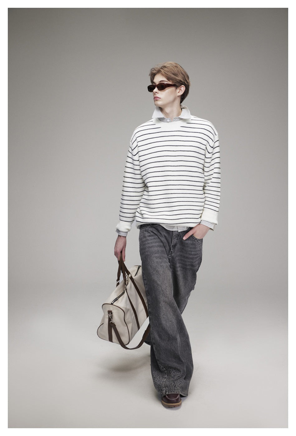 Model wearing white striped knit sweater over a checked shirt with gray jeans, holding a duffel bag