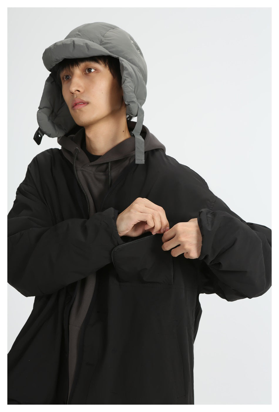 Model wearing a black oversized jacket over a dark hoodie, adjusting its front; gray padded hat with ear flaps.