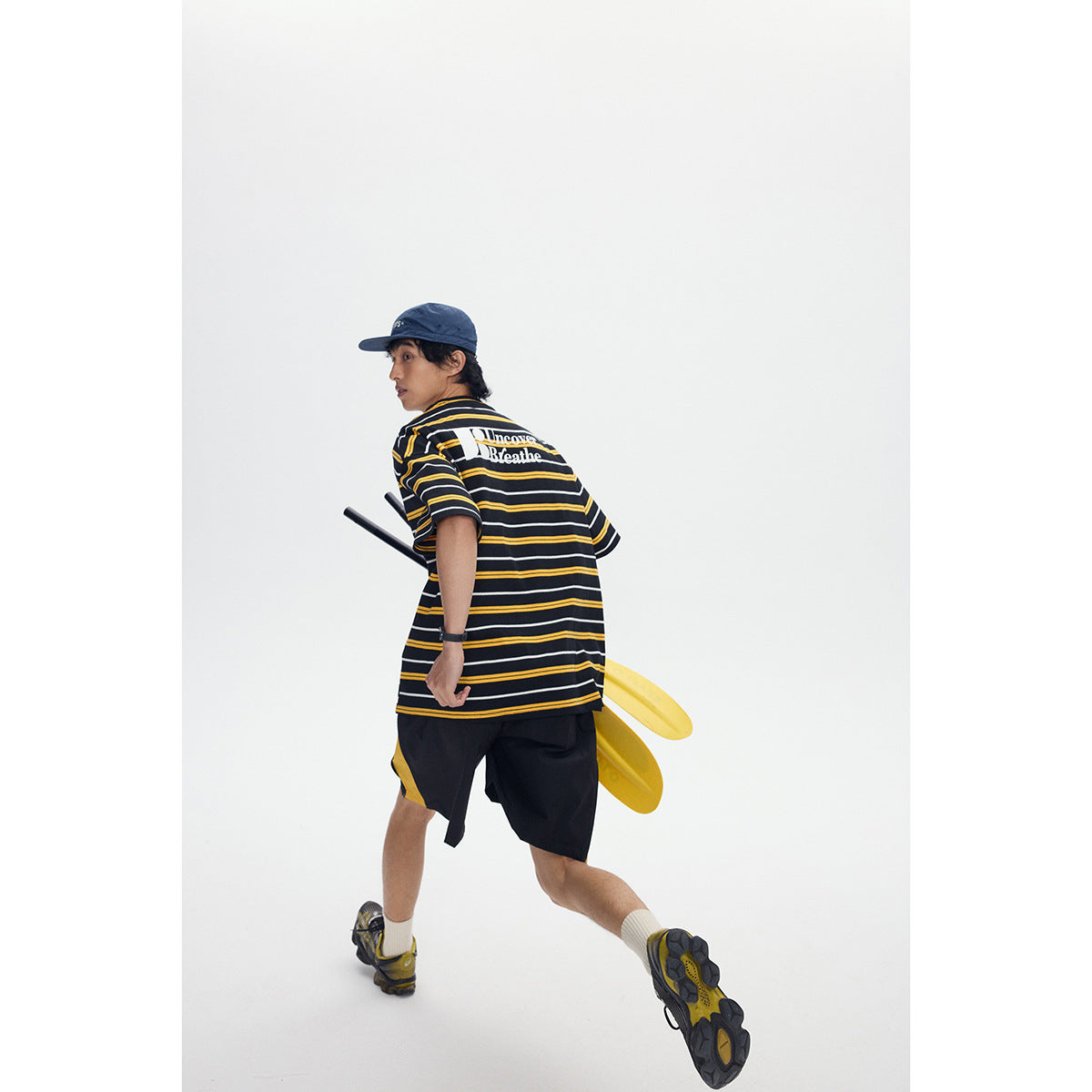 Model turning while wearing the striped black T-shirt with a white back graphic and black shorts with yellow side panels.