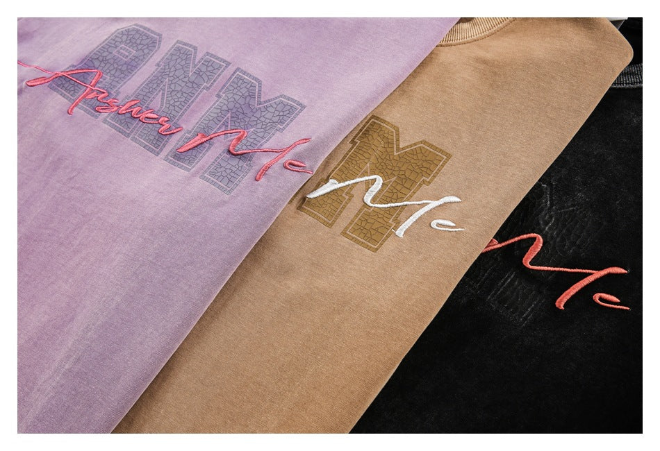 Close-up of purple, tan-brown, and black T-shirts with large textured lettering and cursive embroidery.