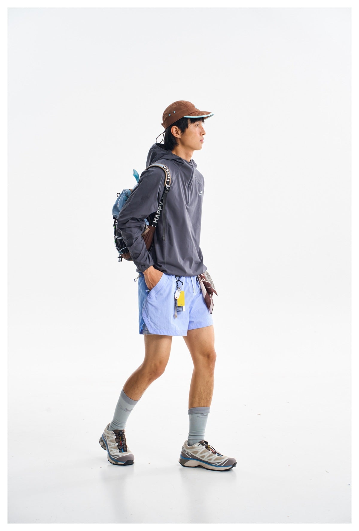 A man wears a charcoal hooded long-sleeve top and light blue shorts with a hiking backpack and brown cap.