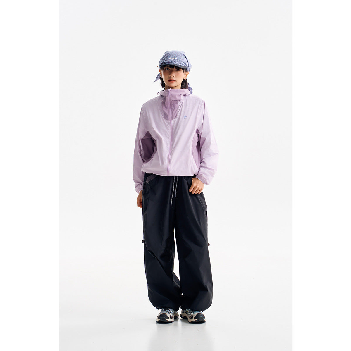 Front view of a person wearing a pale lavender zip jacket, loose black pants, and a blue cap.