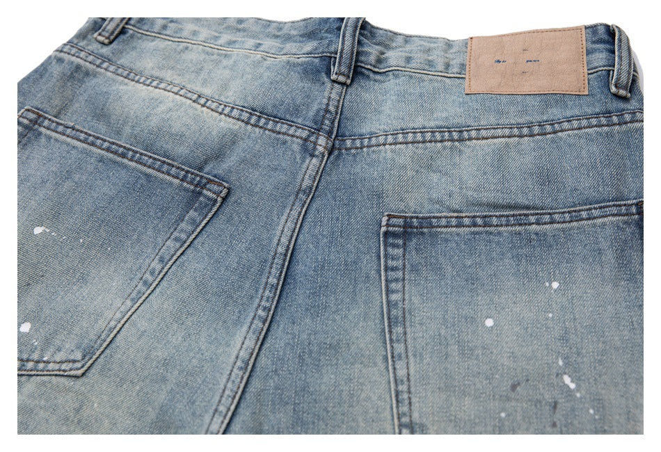 Cropped flat lay of the back of light blue paint-splattered jeans with belt loops, a leather patch, and pockets.