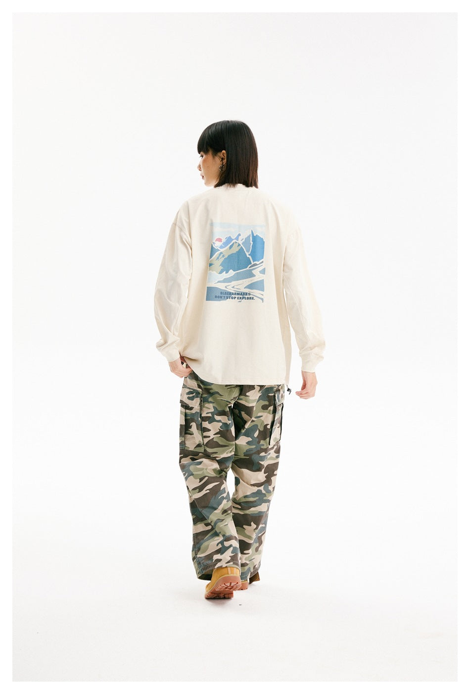 Rear view of a model wearing an ivory long-sleeve shirt with a blue mountain graphic and green camouflage cargo pants.