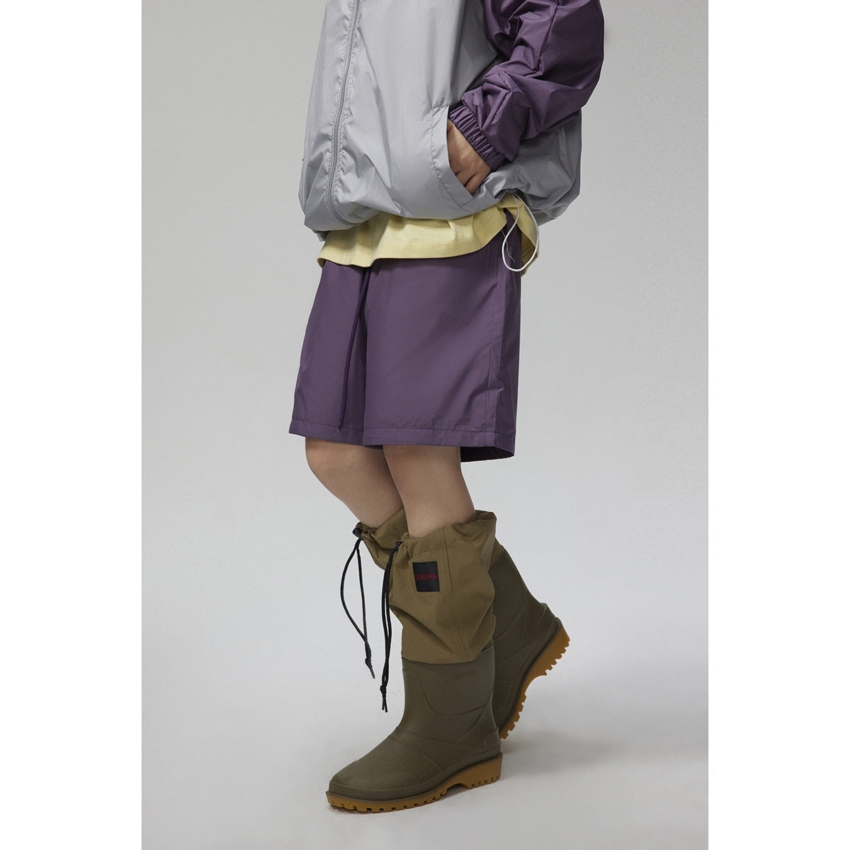 Close-up of purple shorts, a gray-and-purple jacket hem, and olive rain boots with gathered gaiter covers.