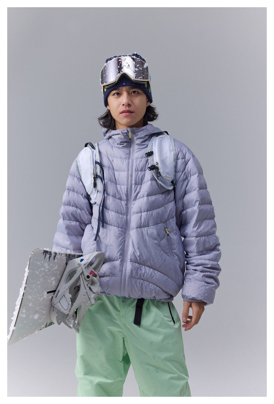 Person wearing a light purple hooded quilted jacket with a backpack, snowboard, goggles and green pants