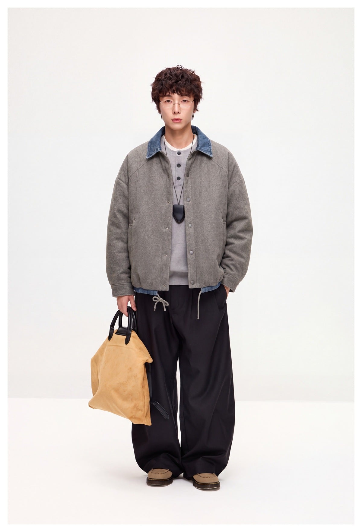 Model wearing gray snap-front jacket with blue denim collar over gray henley, black wide pants, holding tan tote