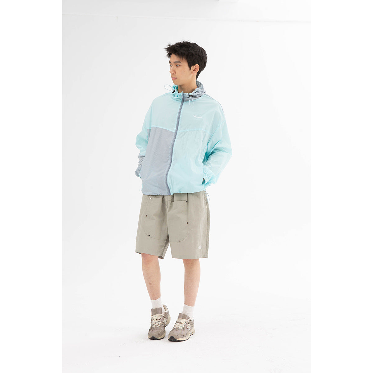 Model wearing a pale aqua and gray hooded jacket with taupe utility shorts, shown full length from the front.
