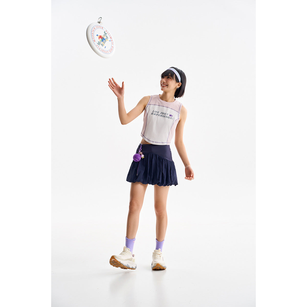 Model wearing a pale pink sleeveless crop top and navy skirt, tossing a round printed object.