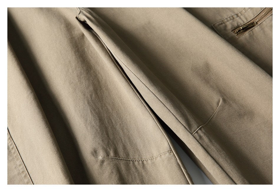 Close-up of taupe trousers showing a stitched seam, overlapping fabric panel, and part of a zipper pocket.