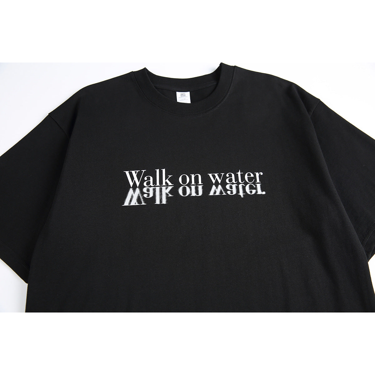 Close view of a black crew-neck T-shirt with white “Walk on water” text and a mirrored reflection graphic.