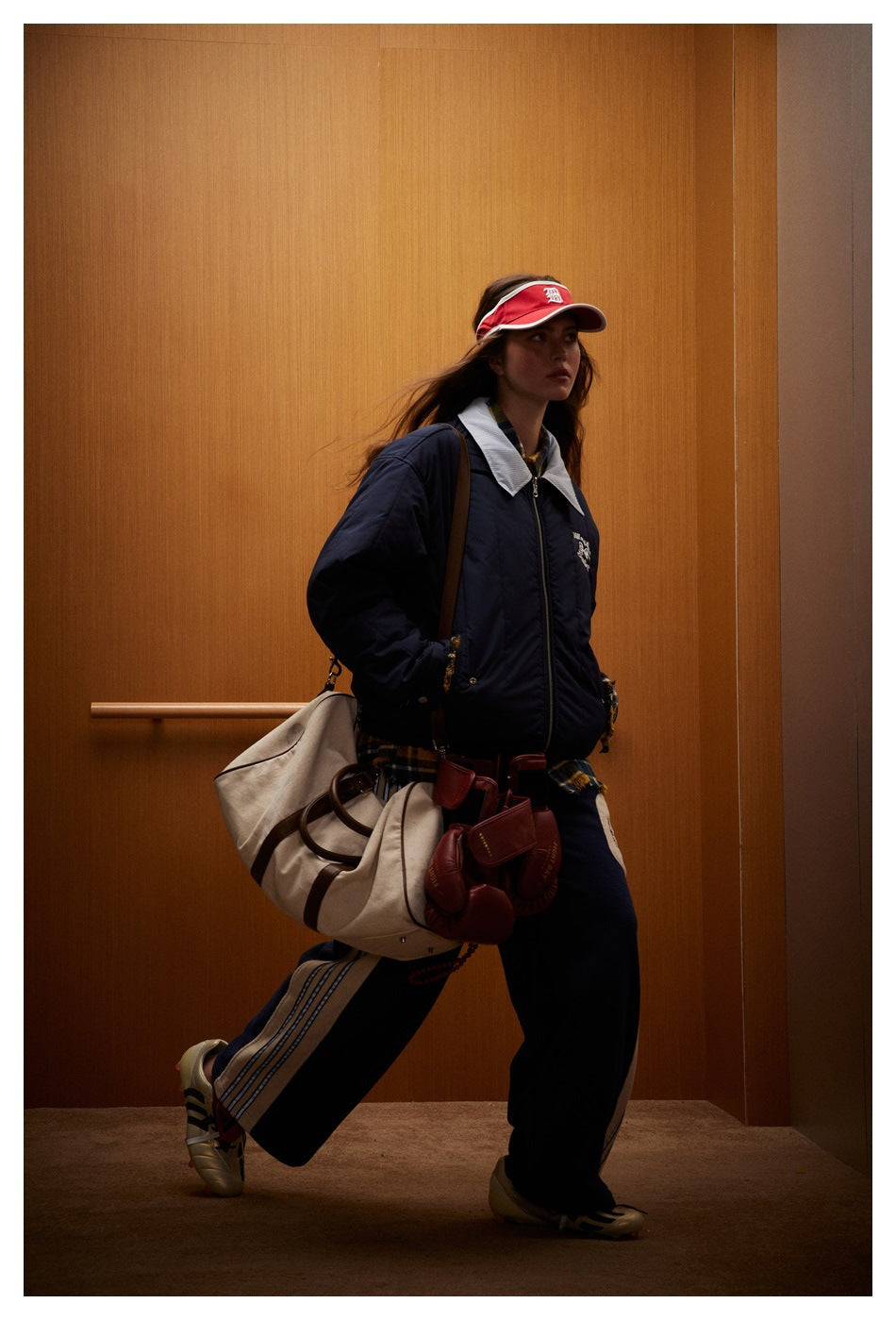Side view of a model wearing a navy jacket and trousers with a light side stripe, carrying a large beige duffel bag.