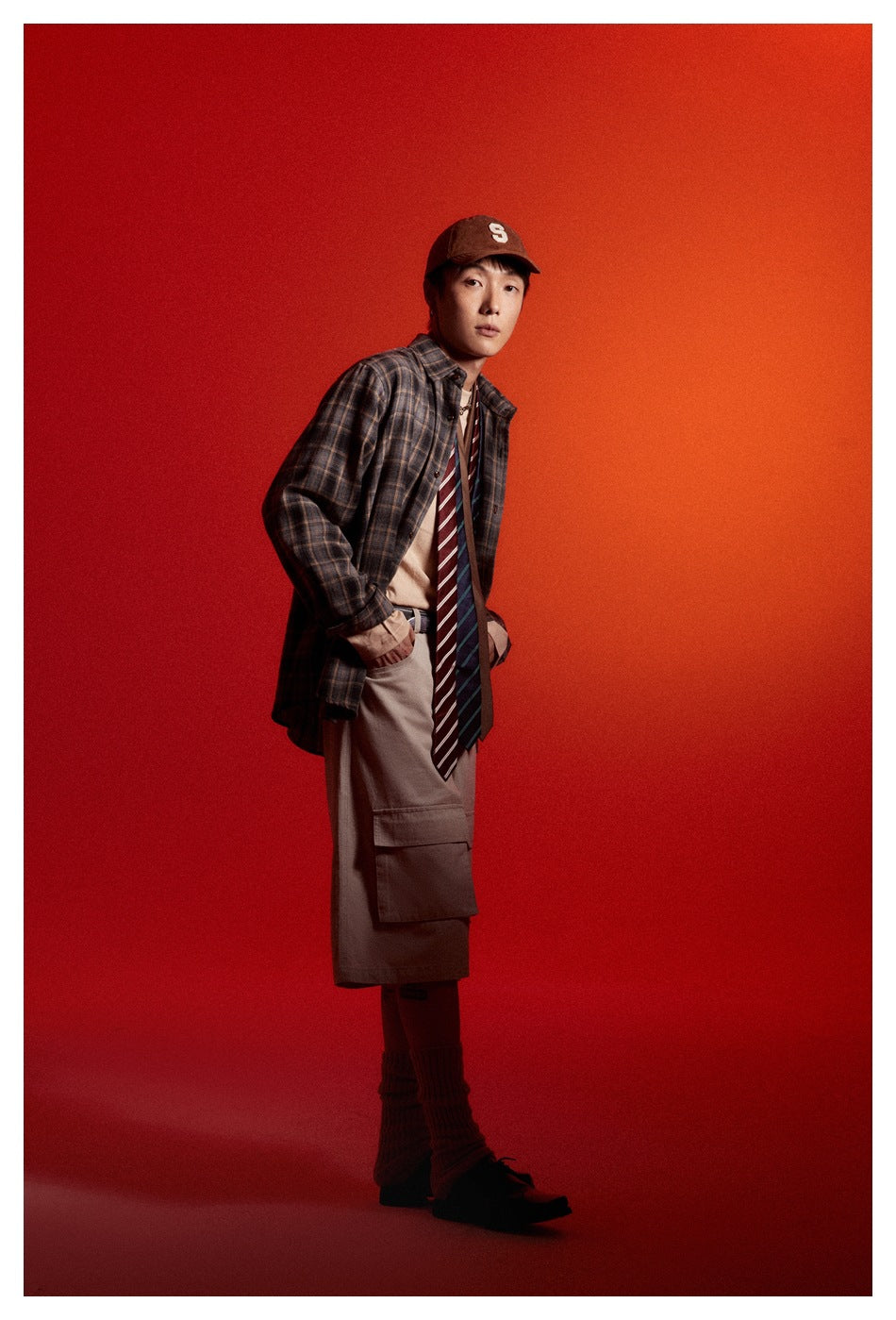 Model in a blue-gray plaid button-up shirt and taupe cargo shorts, shown in a three-quarter pose against red.